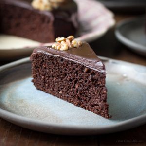 A slice of moist chocolate walnut cake with chocolate ganache and a walnut half served on a blue plate with the larger cake behind.