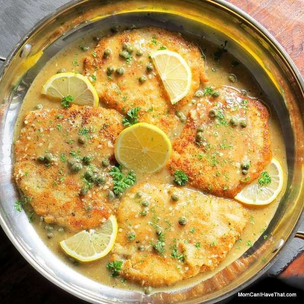 Top down view of 4 low carb breaded pork cutlets in a lemon butter caper sauce garnishes with lemon slices and parsley in a round stainless steel pan.