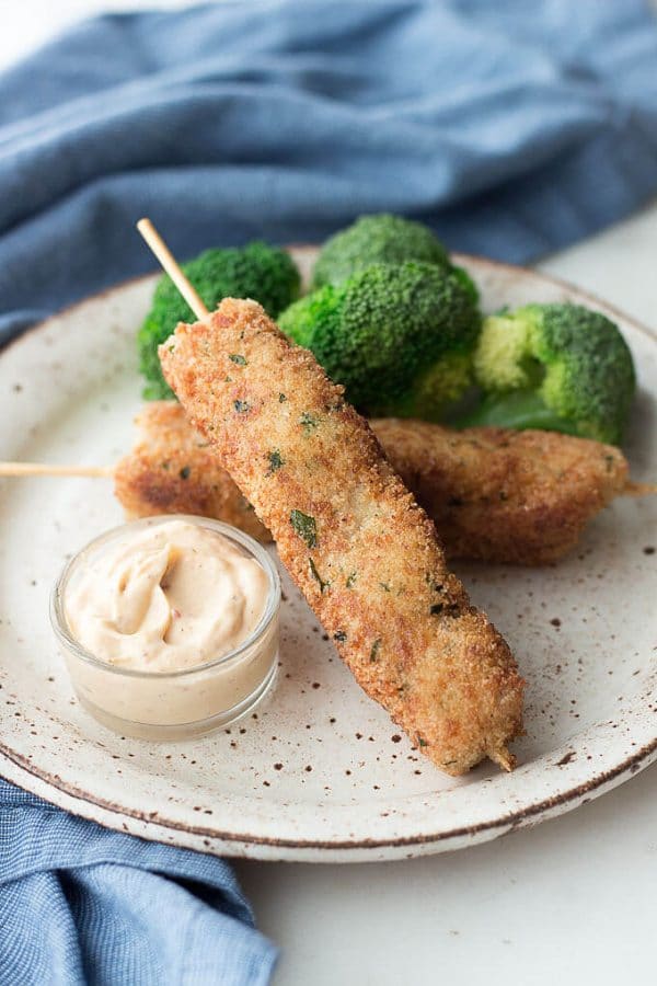 Skewers of city chicken in a low carb gluten free coating on a hand made ceramic speckled plate with dipping sauce to the lower left, steamed broccoli to the upper right and a blue napkin behind.