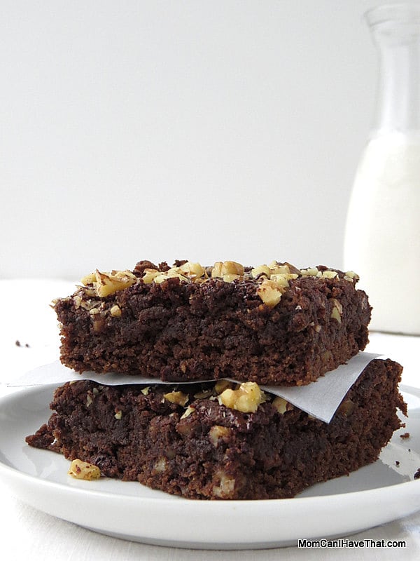 Two low carb chocolate brownies topped with walnuts stacked on a white plate with a glass jar of almond milk in the background. 