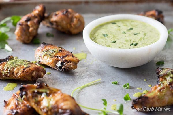 A side view of Indian tikka seasoned grilled chicken wings arranged on a cookie sheet and drizzled with green cilantro sauce from a white marble bowl filled with the sauce.