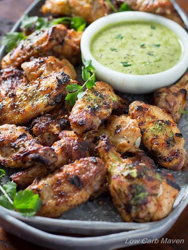 Grilled Indian tikka chicken wings piled around a white bowl of cilantro lime sauce on a gray handmade pottery handled tray placed on a dark wooden table.