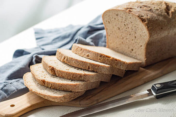 A sliced loaf of low carb bread sitting on a handled cutting board between a knife and a blue napkin.