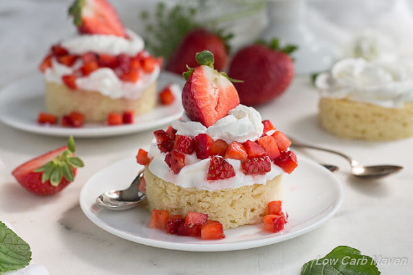 Moist almond flour cake on a round white plate, topped with whipped cream and diced fresh strawberries topped with more whipped cream and a large strawberry half with the leaves still attached. Another strawberry shortcake dessert is in the back ground along with fresh strawberries and mint leaves.