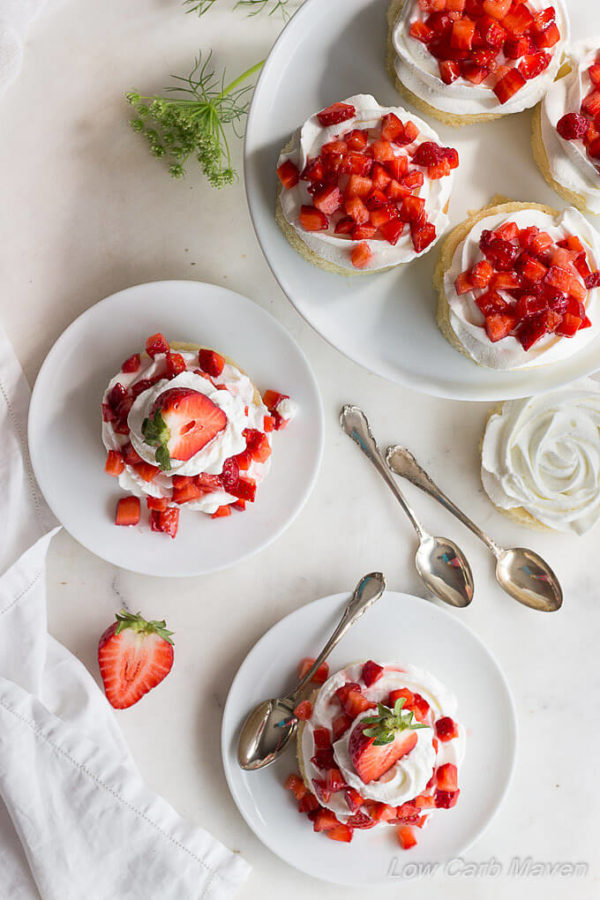 A top down photo of 2 artfully displayed low carb strawberry shortcake desserts on white porcelain plates (on with a spoon) topped with pillows of whipped cream and diced strawberries with a white napkin to the left corner and a raised white plate with 4 partially decorated strawberry desserts in the top right corner.