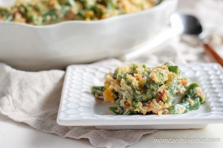 A serving of Keto green bean casserole with crunchy topping on a white square plate with serving dish and spoon in background.
