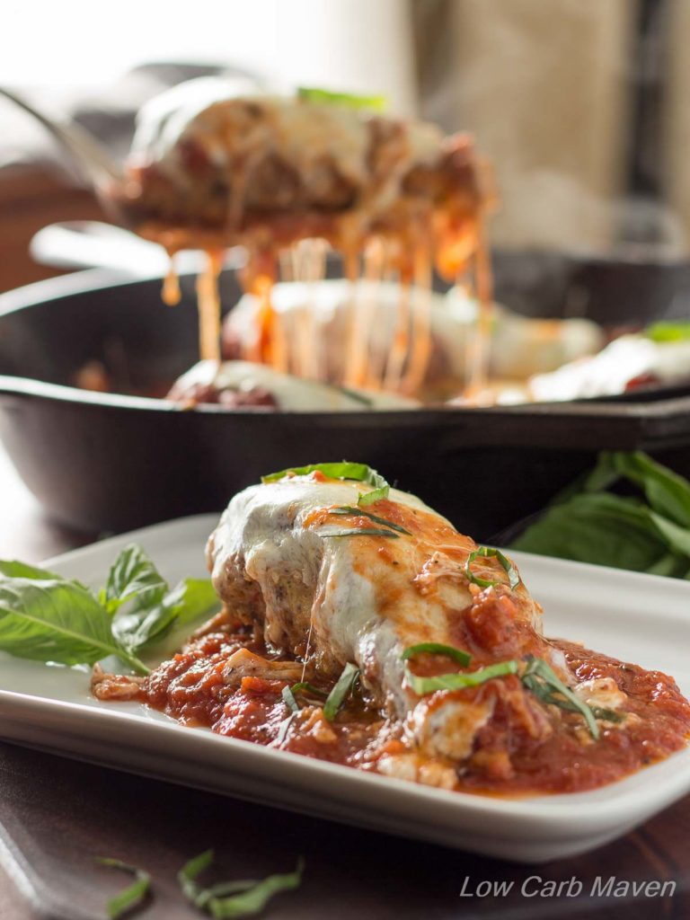 A serving of low carb chicken Parmesan on a plate with melty cheese and garnished with basil. A serving of chicken Parmesan is being lifted by a spatula from a skillet in the background.