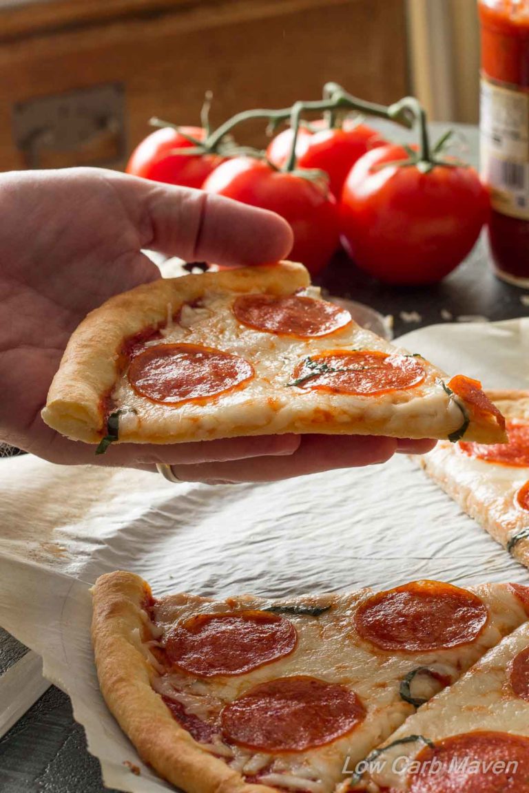 Sliced Low Carb Pepperoni Pizza with a hand held slice and tomatoes in the background.