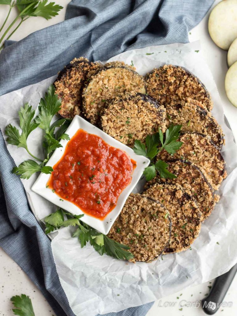 Crispy fried eggplant rounds arranged on a parchment lined plate with parsley garnish and marinara sauce with a blue napkin tucked under the plate to the left and a knife and cut raw eggplant rounds to the right.