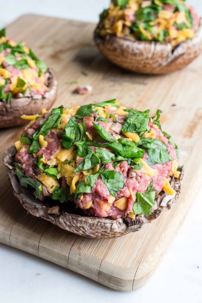 Uncooked stuffed portobello mushrooms with ground beef,spinach and cheese sitting on a cutting board.