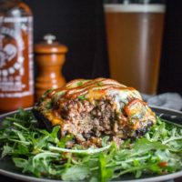 Portobello mushroom stuffed with ground beef, spinach, and cheese on an arugula salad with beer and siracha hot sauce in the background.