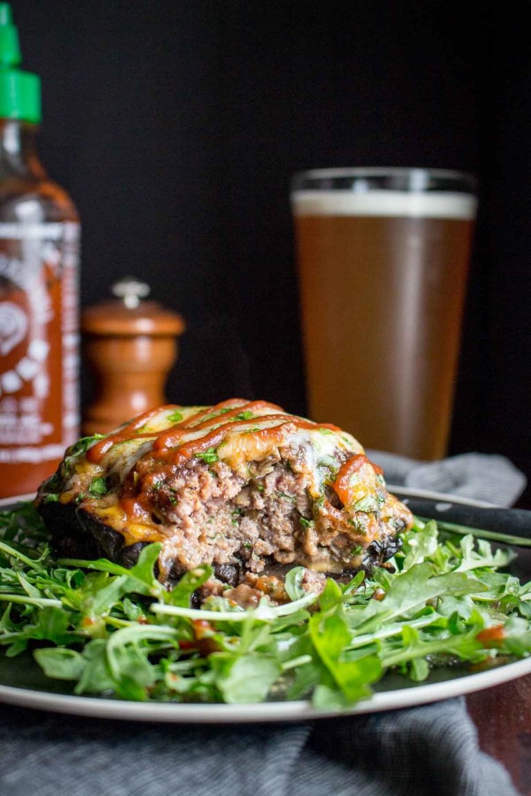 Portobello mushroom stuffed with ground beef, spinach, and cheese on an arugula salad with beer and siracha hot sauce in the background.