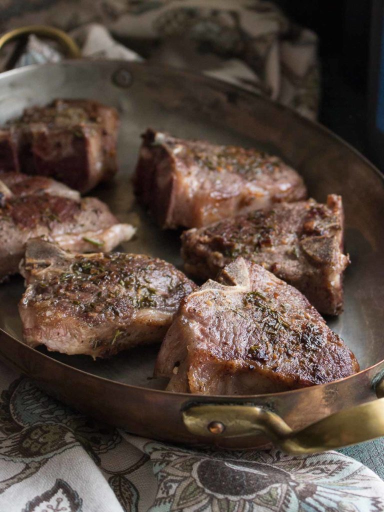 Pan seared lamb chops with rosemary and garlic in an oval copper baking pan with brass handles on a floral decorative napkin.