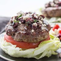 Bun-less lamb burger with lettuce, tomato, Greek olives, feta cheese and mint on a plate.