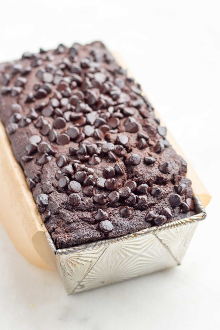 Top view of a baked loaf of low carb chocolate pound cake topped with chocolate chips in a silver vintage starburst pattern Ovenex loaf pan with brown parchment paper over-hanging the sides. 
