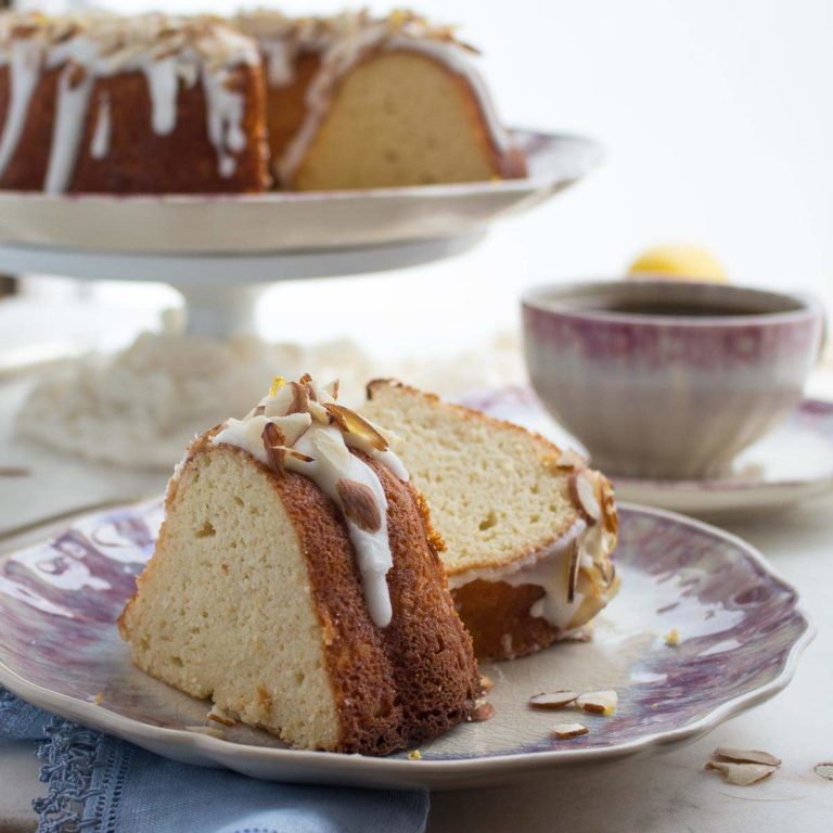 Two slices of almond flour bundt cake drizzled with lemon glaze and topped with almonds on a purple plate with a cup of coffee and the larger cake behind.