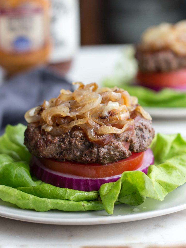 Low carb bunless burger on tomato, red onion and lettuce on a plate topped with BBQ sauce and caramelized onions.