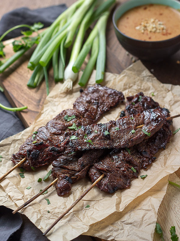 Skewered and grilled Beef Satay on parchment on a cutting board with scallions and a bowl of peanut sauce.