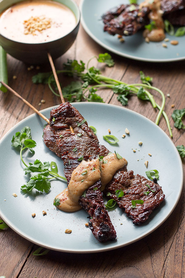 Beef Satay on skewers with Peanut Sauce on a blue plate with cilantro with peanut sauce in a bowl behind.