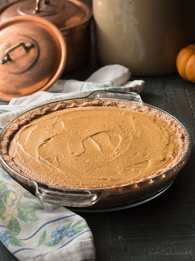 Whole sugar-free pumpkin pie with a nut crust in a glass pie plate with a floral napkin to the left and copper pots in the back ground.