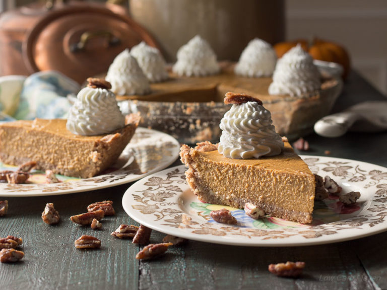 Two slices of sugar-free pumpkin pie with whipped cream on china plates with the remaining pumpkin pie in a pie plate behind. Pecans are scattered on the wooden surface in the foreground.