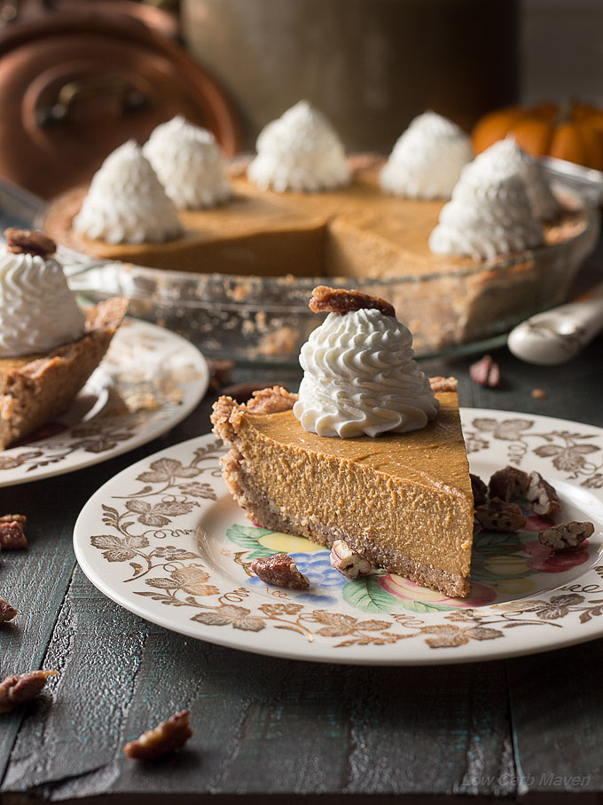 A slice of sugar-free pumpkin pie with a large ruffled dollop of whipped cream on a china plate decorated with gold grape leaves. A sliced pie is in the background decorated with mountains of piped ruffled whipped cream.