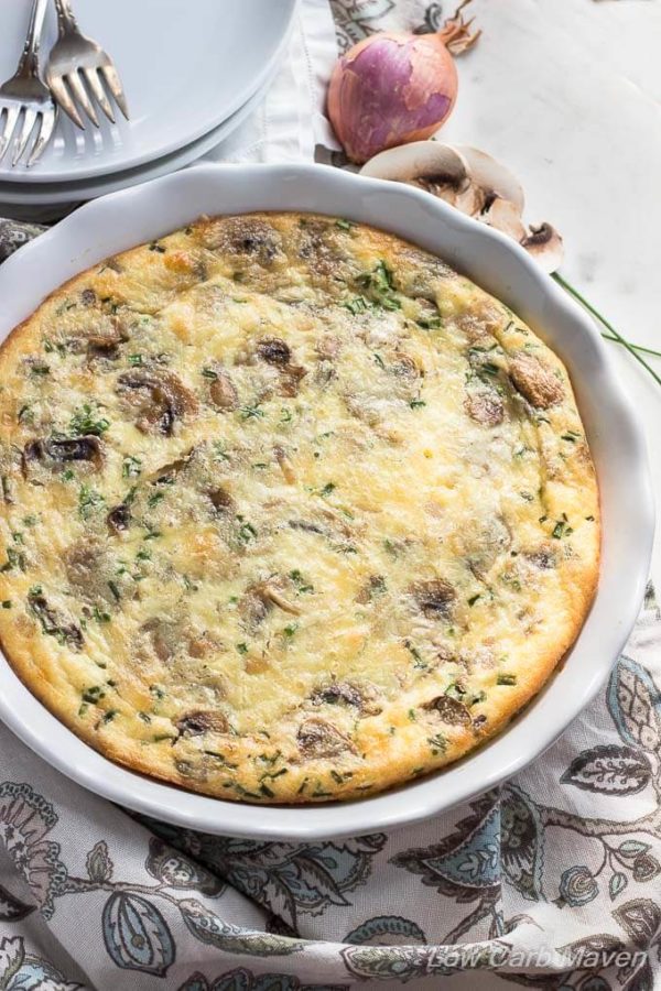 A top down view of a crustless mushroom quiche in a white fluted pie dish with a floral napkin arranged to the bottom of the dish, white plates with forks in the upper left corner and a shallot and sliced mushrooms to the upper right.