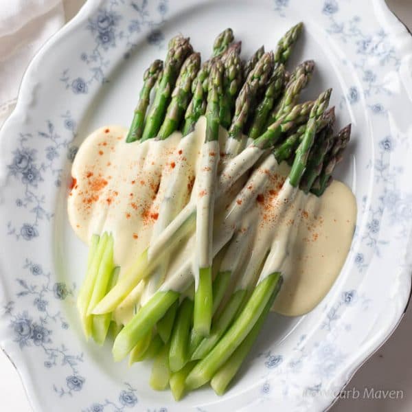 A top-down view of bright green steamed asparagus spears dressed with a rich yellow sauce of browned butter Hollandaise sprinkled with paprika arranged on a oval blue and white floral china plate.