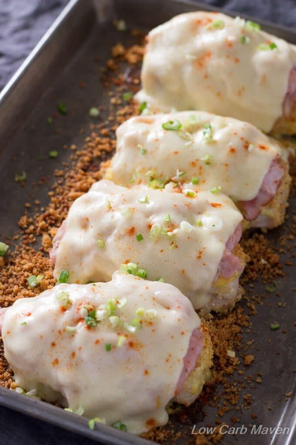 Four baked chicken breasts topped with low carb breading, slices of ham and melted Swiss cheese with sliced scallions on top in a small metal sheet pan with additional toasted crumbs in the bottom of the pan.