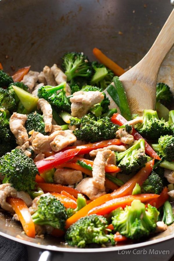 A bamboo spatula stirring a pork stir-fry of sliced pork, red bell peppers, orange bell peppers, scallions and broccoli in a carbon steel wok.