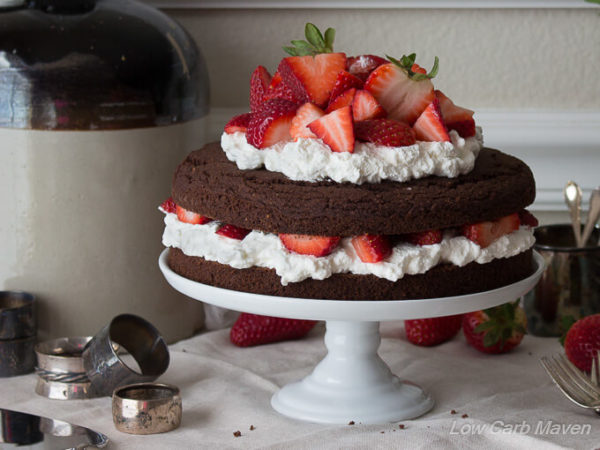 Chocolate cake on a white pedestal filled and topped with pillows of whipped cream and sliced fresh strawberries on a cloth covered surface with antique silver napkin rings and crockery to the left and behind and strawberries and silver forks to the right.