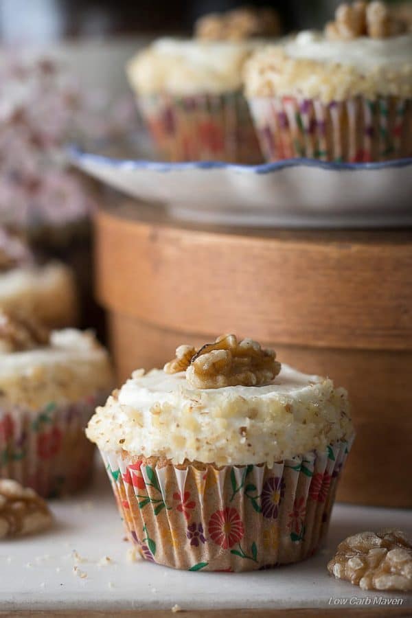 Low carb sugar free carrot cake muffin with fluffy cream cheese frosting decorated with finely chopped walnuts around the side and a walnut half on top in a colorful floral fluted cupcake paper with more cupcakes to the side fading into the background and two cupcakes sitting on a blue and white ruffled plate on a brown round wooden cheese box behind.