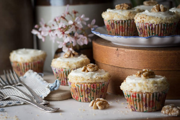 Four sugar free carrot cake cupcakes with cream cheese frosting, decorated with walnuts and colorful floral cupcake papers sitting on a white marble surface between 3 silver forks on a vintage blue and white crocheted pot holder and a blue and white plate of cupcakes sitting on a round cheese box with pink flowers and vintage crockery in the background.