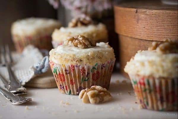 Close up of carrot cake cupcakes in colorful floral cupcake papers decorated with fluffy cream cheese frosting, finely chopped walnuts, and walnut halves with forks on a white cloth to the left and a brown wooden round box to the right.