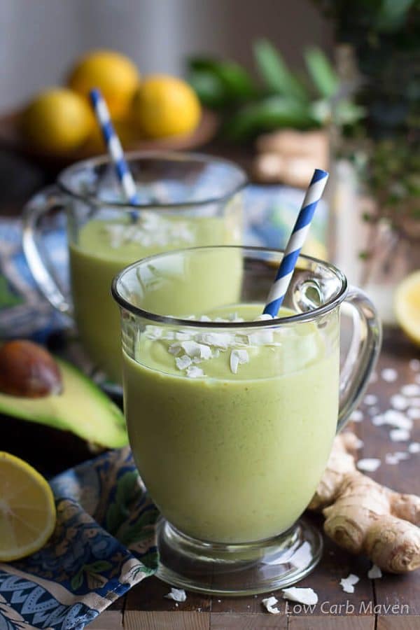 Two green smoothies, one in front of the other, in clear handled. footed mugs with blue and white striped straws. next to a colorful blue napkin with cut avocados, lemons and ginger.
