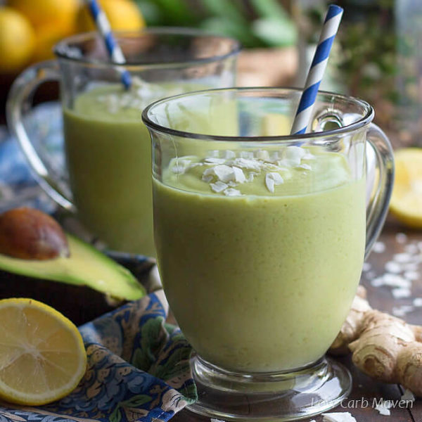 Close up of a green avocado coconut milk smoothie in a clear glass mug with flaked coconut and a straw next to a blue napkin with cut lemons, avocado and ginger.