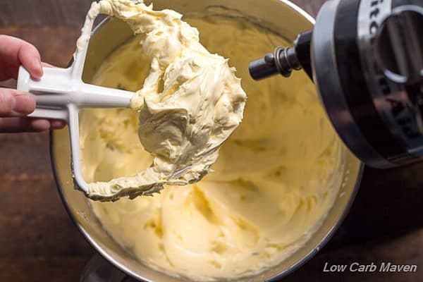 Rich, buttery and silky sugar free French buttercream on a white Kitchen Aid mixer paddle being held over a stainless steel bowl of whipped yellow french buttercream with part of the black mixer showing on the right.
