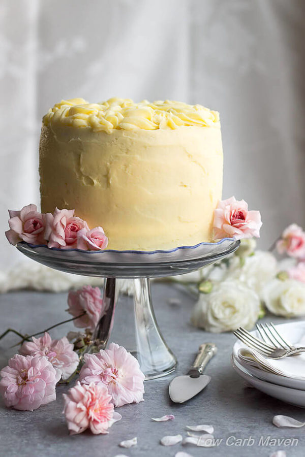 A layer cake decorated in yellow frosting with rosettes on top sitting on a clear pedestal with white and pink roses.