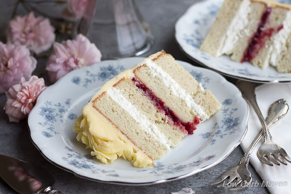 A slice of white layer cake filled with raspberry jam and whipped cream, decorated in yellow French buttercream frosting on a blue and white china plate with pink roses and silver forks. 