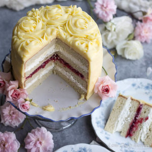 Cut white layer cake on a pedestal decorated with yellow frosting and rosettes on a table decorated with white and pink roses and blue and white china dishes.