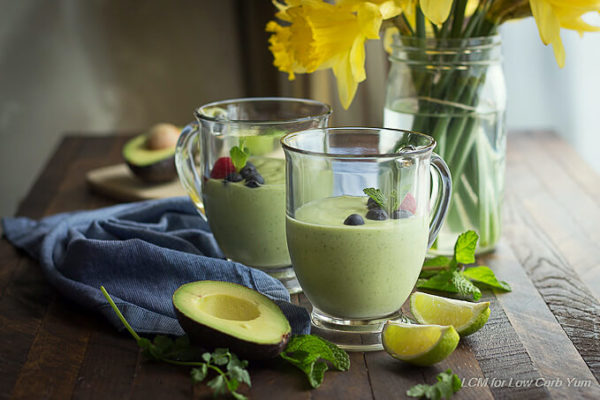 Green avocado smoothies garnished with berries and mint in glass handled mugs with lime, mint and cilantro placed next to a blue napkin and yellow daffodils. 