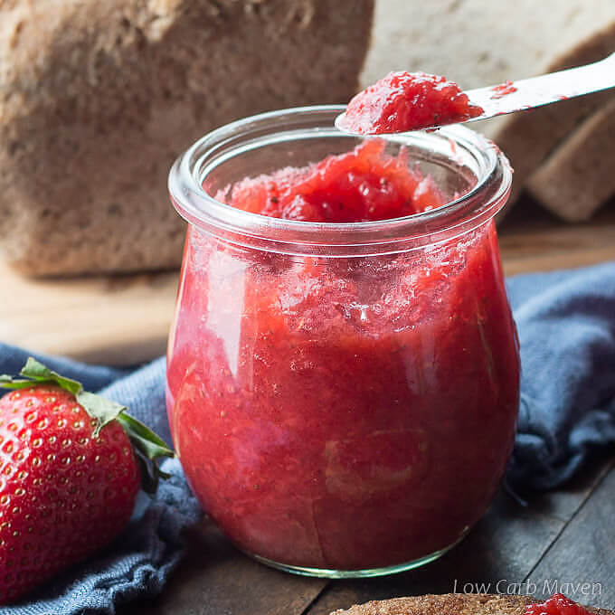 A knife with strawberry jelly on a jar of opened jelly in front of a blue napkin and a sliced loaf of bread on a cutting board.