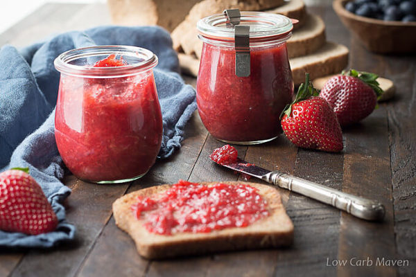 A piece of toast with strawberry jam on a wooden surface with a knife, jars of strawberry jam, strawberries and sliced bread in the background.