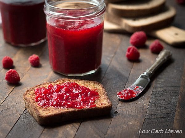 A piece of toasted low carb flax bread spread with raspberry jam and fresh raspberries on a dark brown wooden lath board with a jar of raspberry jam and a silver knife with jam behind. 