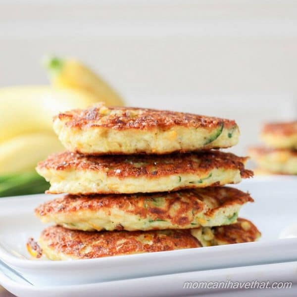 Stacked low carb zucchini fritters on a white plate with yellow squash behind.