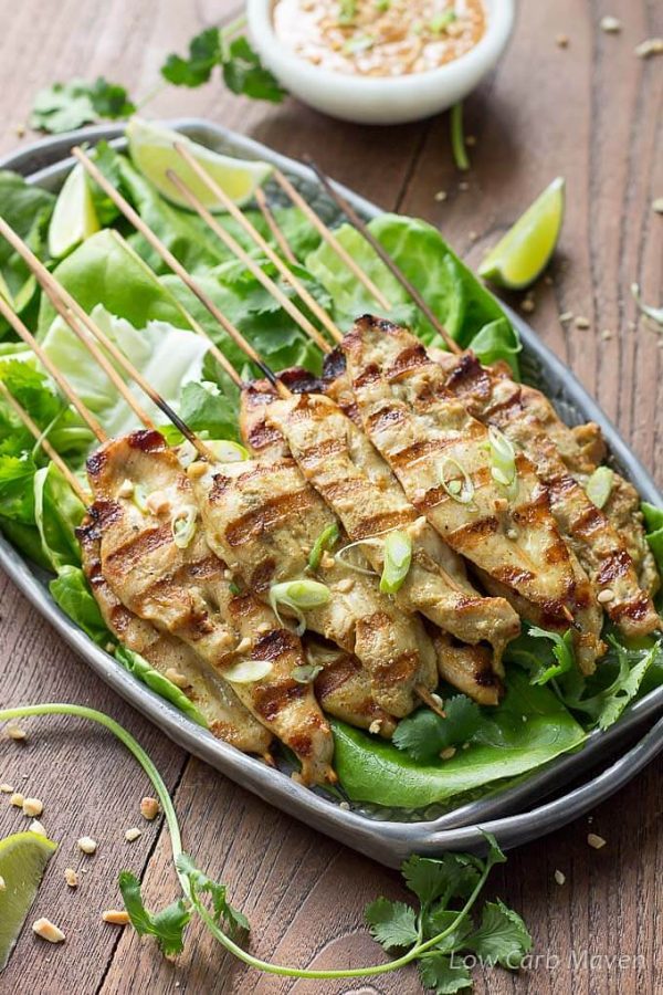 Grilled Thai chicken satay threaded on bamboo skewers on lettuce leaves on a gray plate served with peanut sauce and limes.