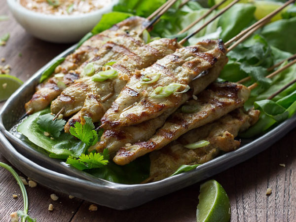 Thai chicken satay, marinated chicken threaded on bamboo skewers and grilled, on a gray serving plate served with peanut sauce and limes.