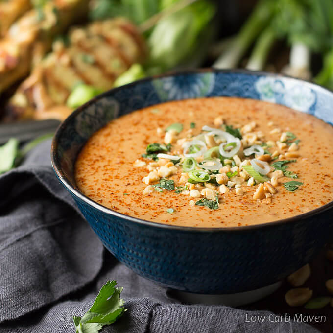 Peanut sauce garnished with cayenne pepper, cilantro, green onion and crushed peanuts in a blue Asian print bowl placed on a dark gray napkin with chicken satay on skewers, scallions and peanuts in the background.