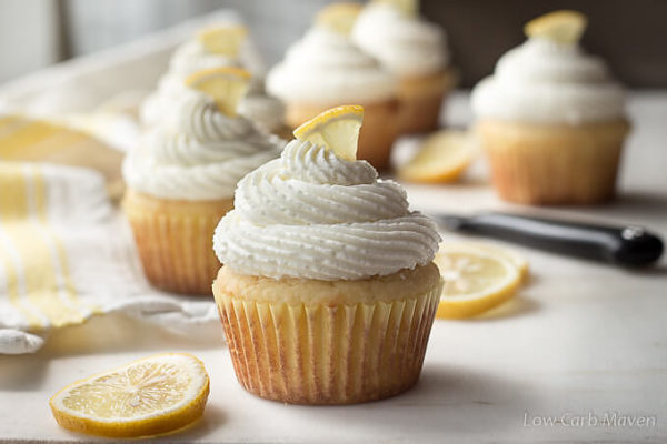 Six lemon cupcakes with cream cheese frosting on a marble surface with sliced lemons and a yellow striped tea towel. 