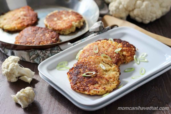 Cauliflower fritters or low carb hash browns stacked off-set on a white rectangular plates, garnished with green onions, and a skillet with more fritters behind with pieces of raw cauliflower in the background.